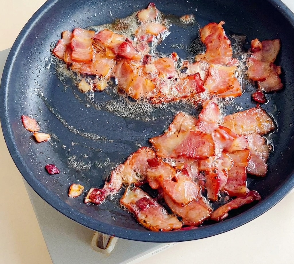 Bacon pieces frying in a pan with onions until browned and crispy.