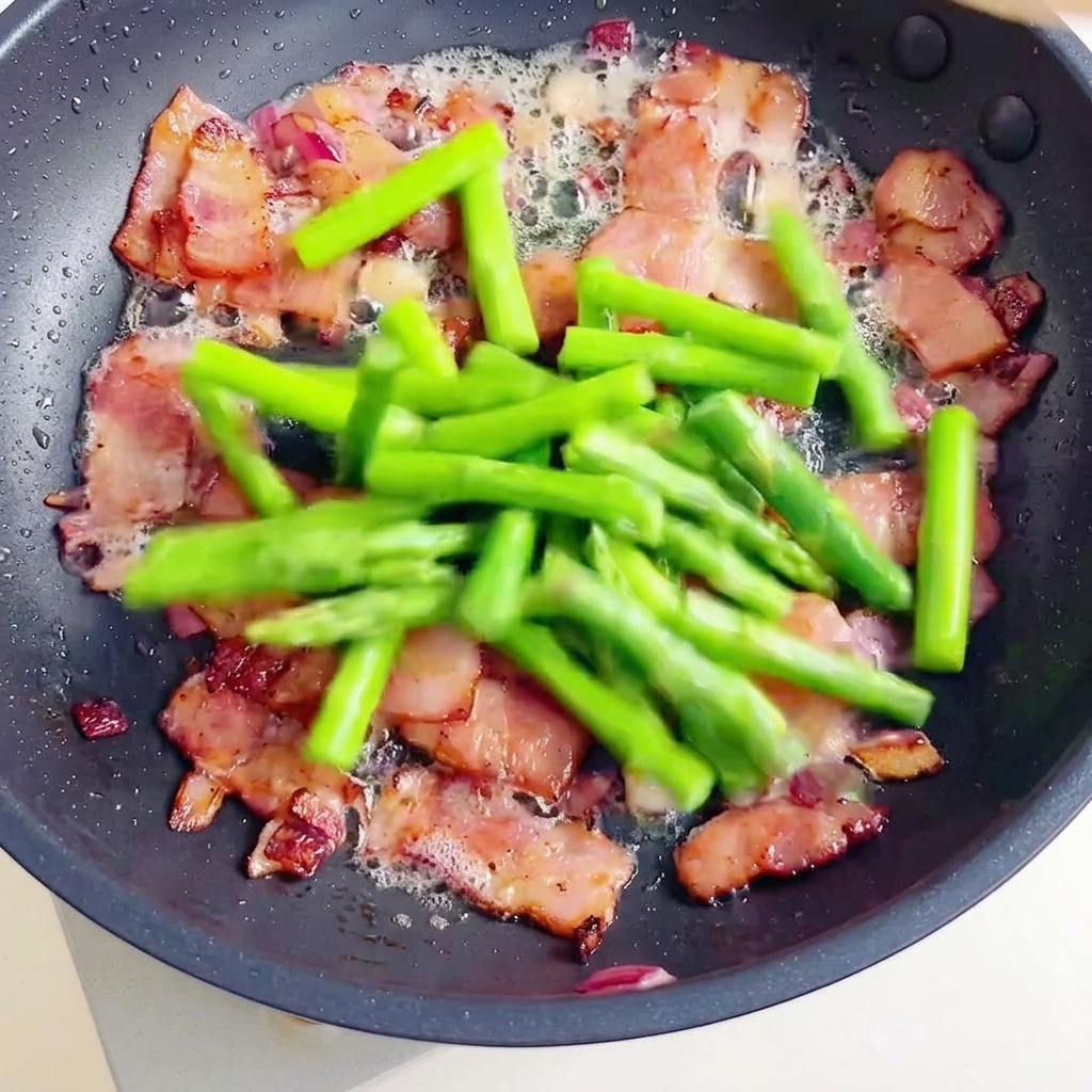 A pan containing crispy bacon pieces and fresh green asparagus segments being joined by cooked spiral fusilli pasta.