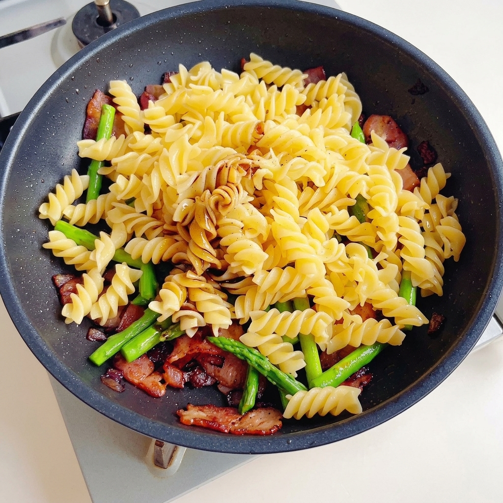 Soy sauce, dark soy sauce, and oyster sauce being poured over a mixture of pasta, bacon, and asparagus in a non-stick pan.