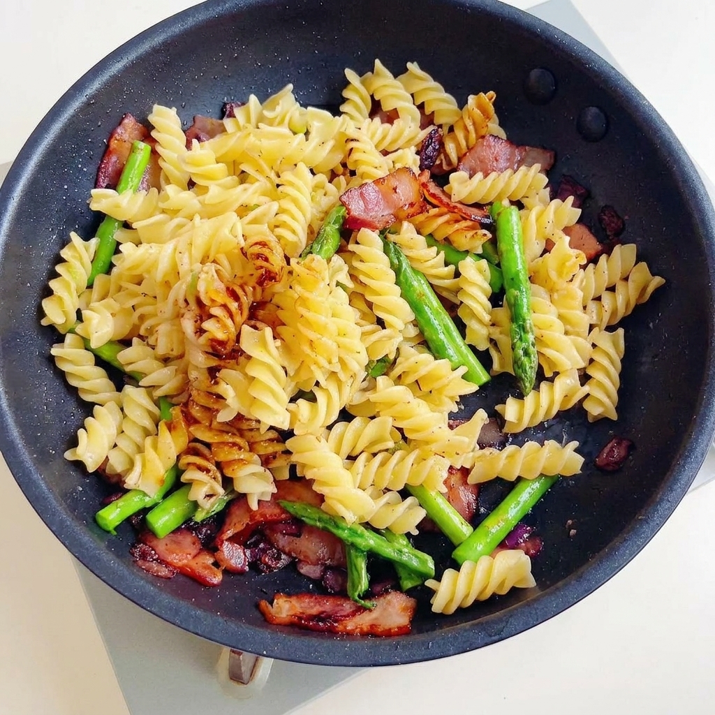 A delicious stir-fry of fusilli pasta, crispy bacon, and asparagus being tossed in a dark, savory sauce in a frying pan.