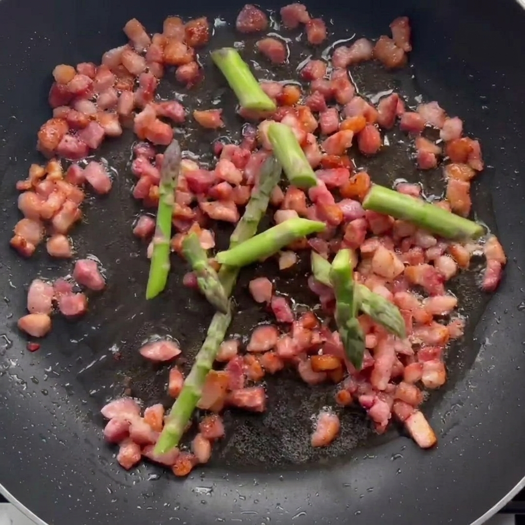 Asparagus pieces cooking in a pan with browned bacon bits.