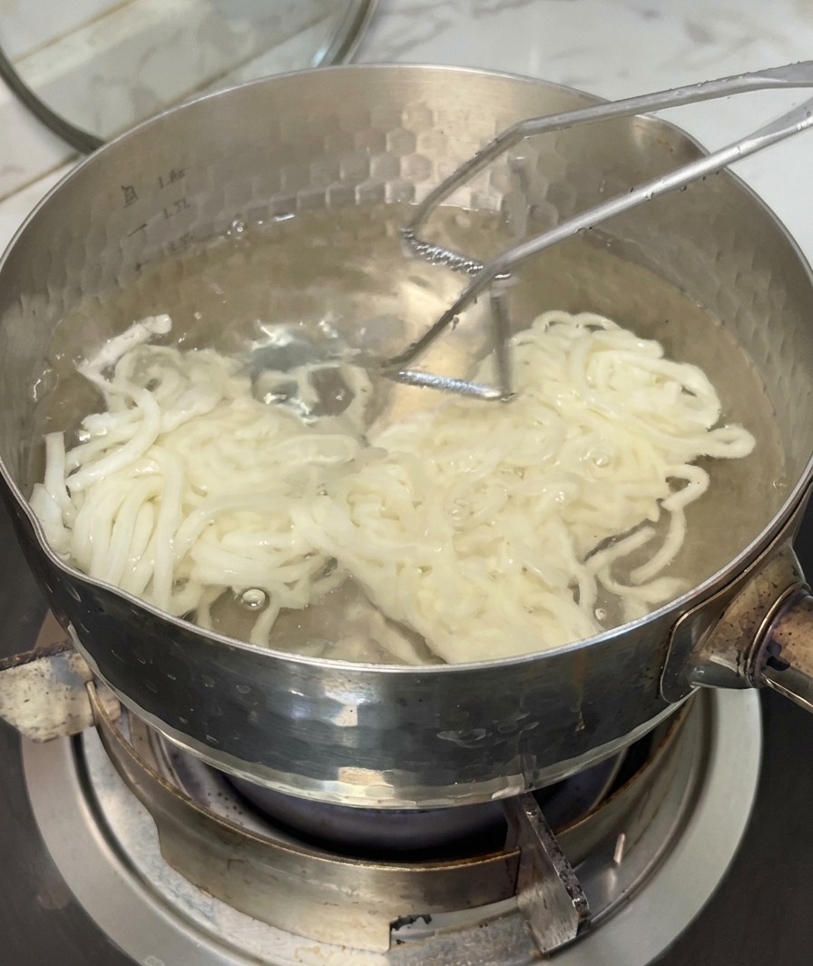 Chewy udon noodles being lifted from a pot of boiling water with a metal strainer.