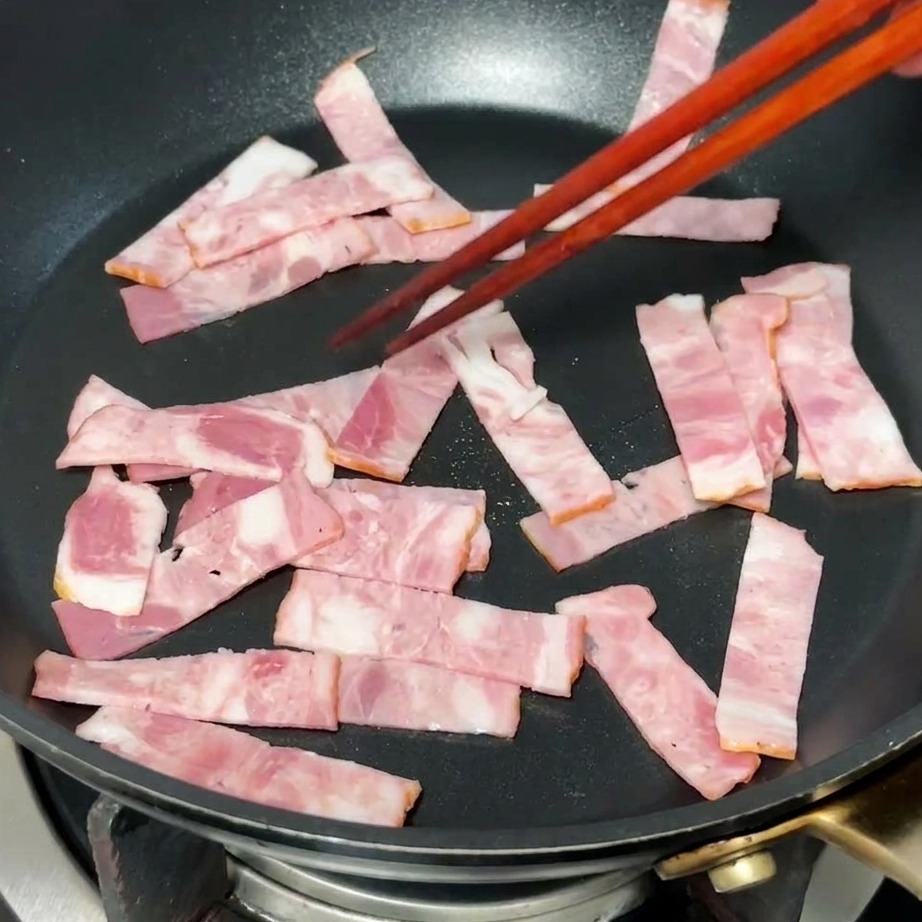 Bacon pieces browning in a hot frying pan, with rendered fat visible.