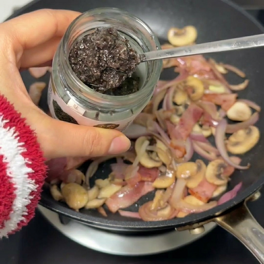 A spoonful of dark, earthy truffle sauce being mixed into a pan containing sautéed bacon, sliced red onions, and mushrooms.