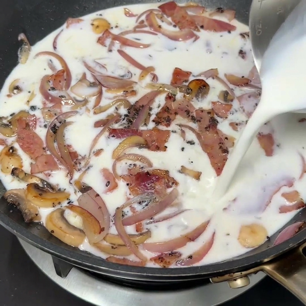Milk being poured from a measuring cup into a pan with stir-fried bacon, onions, and mushrooms.