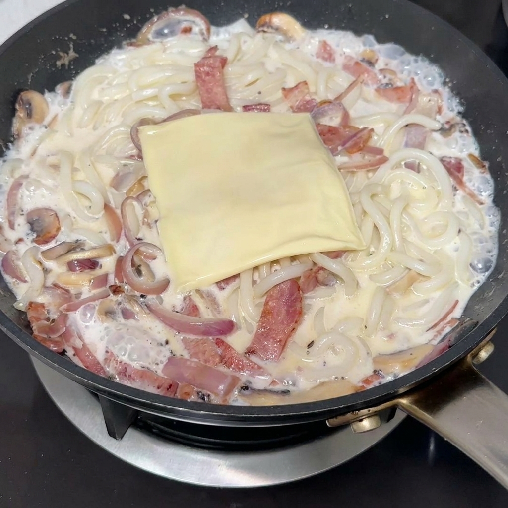 A square slice of processed cheese resting on top of creamy udon noodles and bacon in a frying pan.