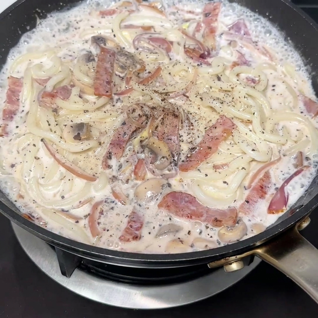 Close-up of freshly cracked black pepper being seasoned over a pan of creamy bacon and mushroom udon noodles.
