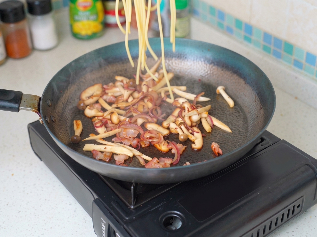 Cooked angel hair pasta being added into a pan filled with sautéed bacon, onions, and mushrooms.