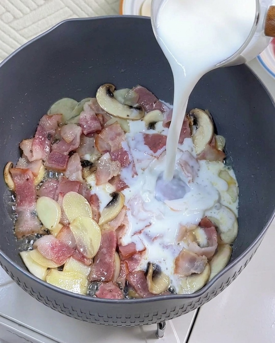 Milk being poured into a pan with sautéed bacon and mushrooms.
