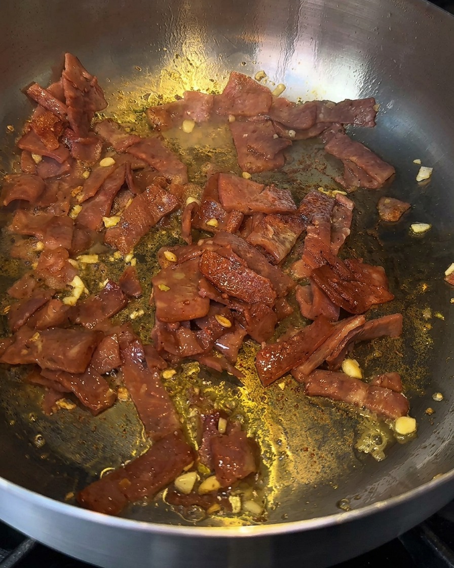 Sizzling bacon and garlic in a pan being deglazed with a splash of cooking liquor.