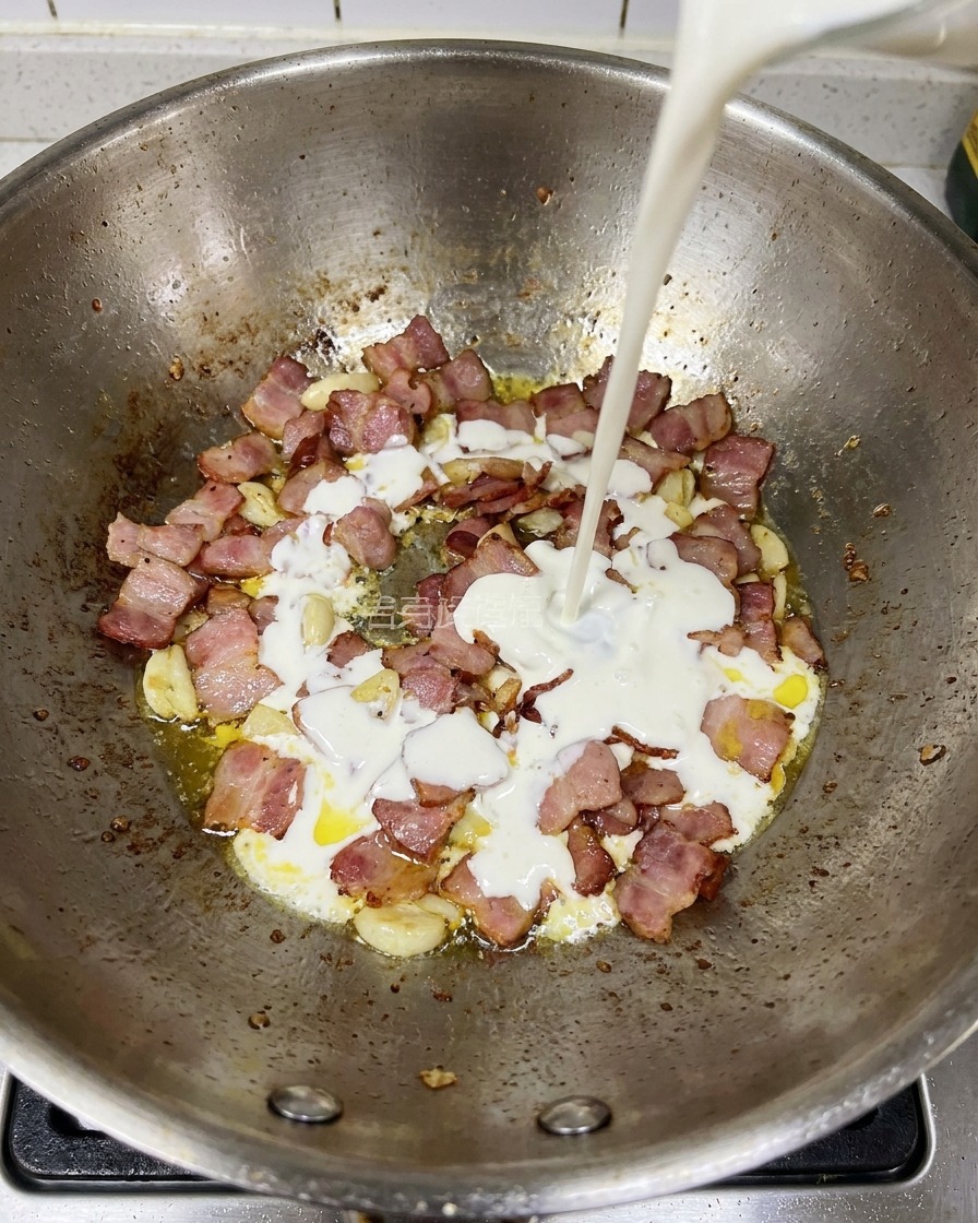 Heavy cream being poured into a pan containing fried bacon and garlic pieces.