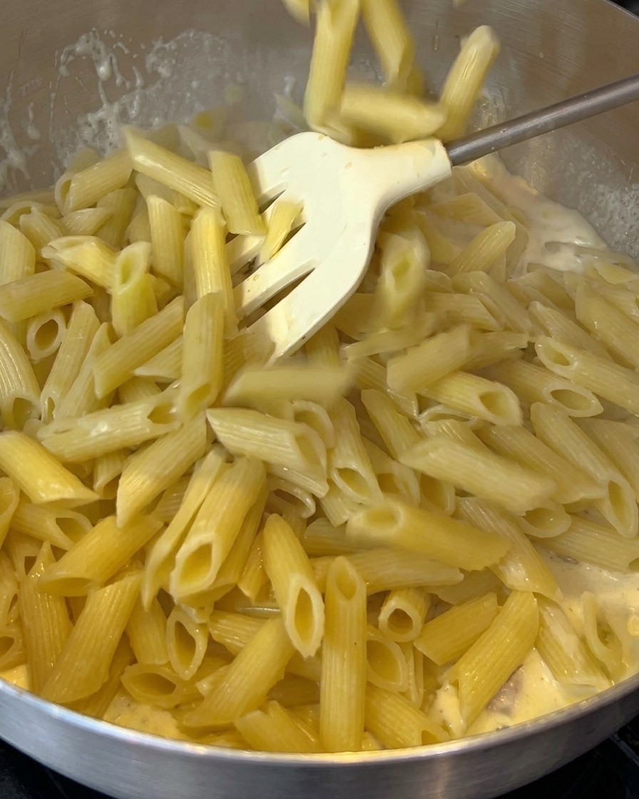 Cooked penne pasta being added to a pan with a creamy bacon sauce.