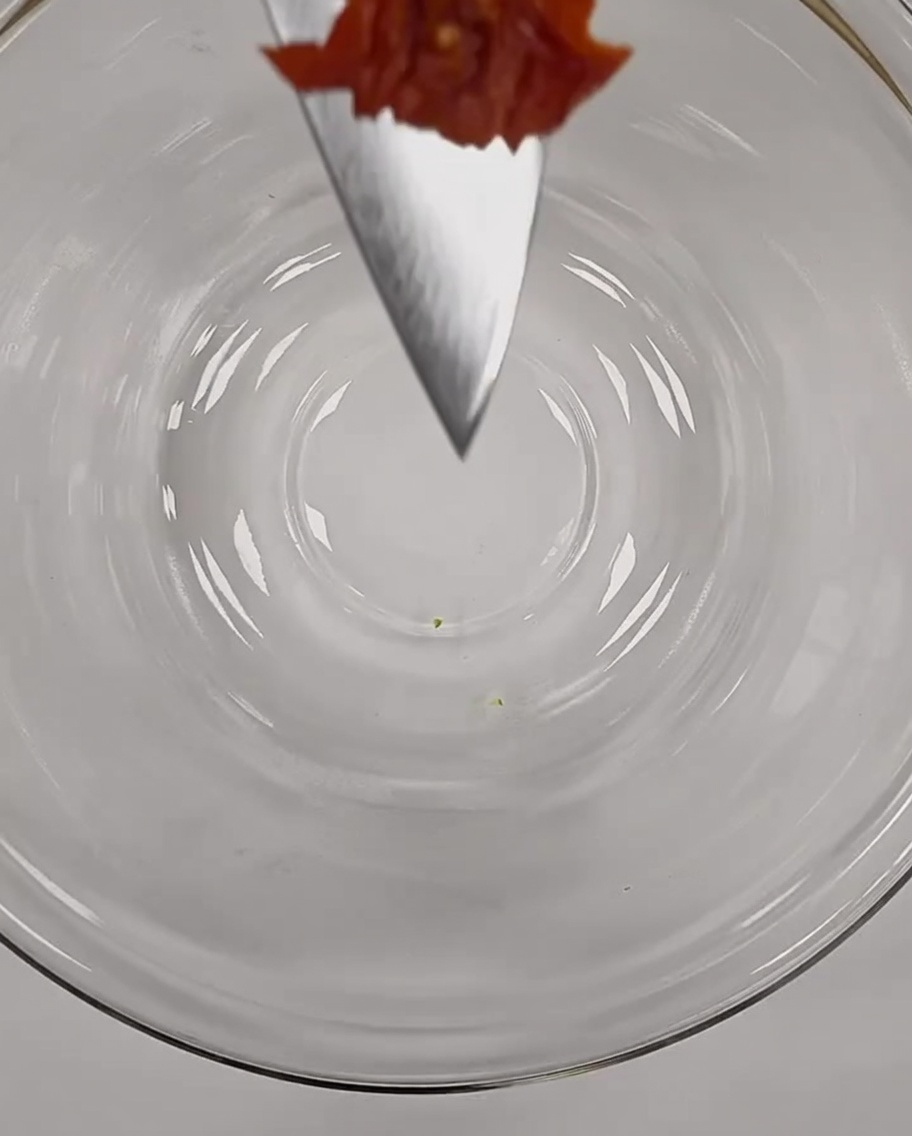 A small, finely chopped piece of red sun-dried tomato delicately balanced on the tip of a chefs knife over a glass bowl.