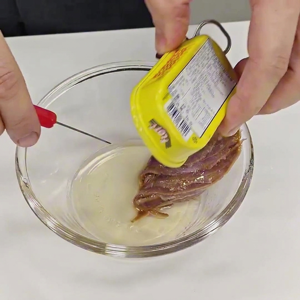 Hands carefully pulling an anchovy fillet from a yellow tin and placing it into a small clear glass bowl.