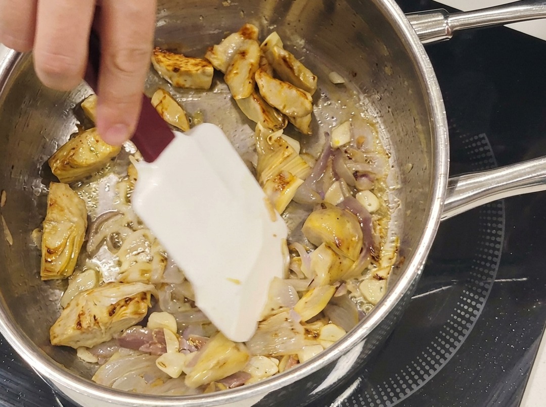 Sautéed artichoke hearts, garlic slices, and anchovies cooking together in a stainless steel pan.