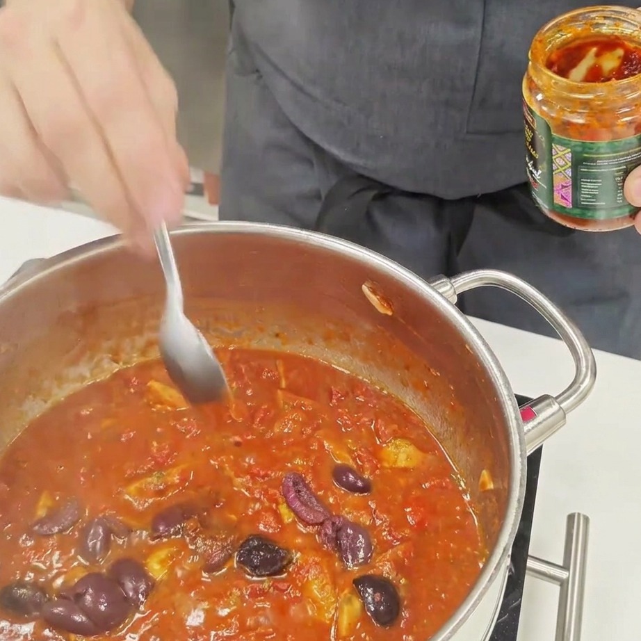 A spoonful of chili sauce being stirred into a bubbling tomato-based pasta sauce.