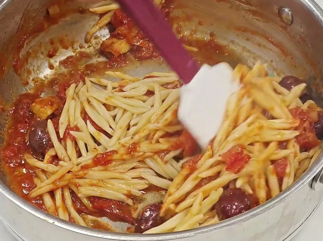Drained Trofie pasta being added to a pan filled with rich red Puttanesca sauce.