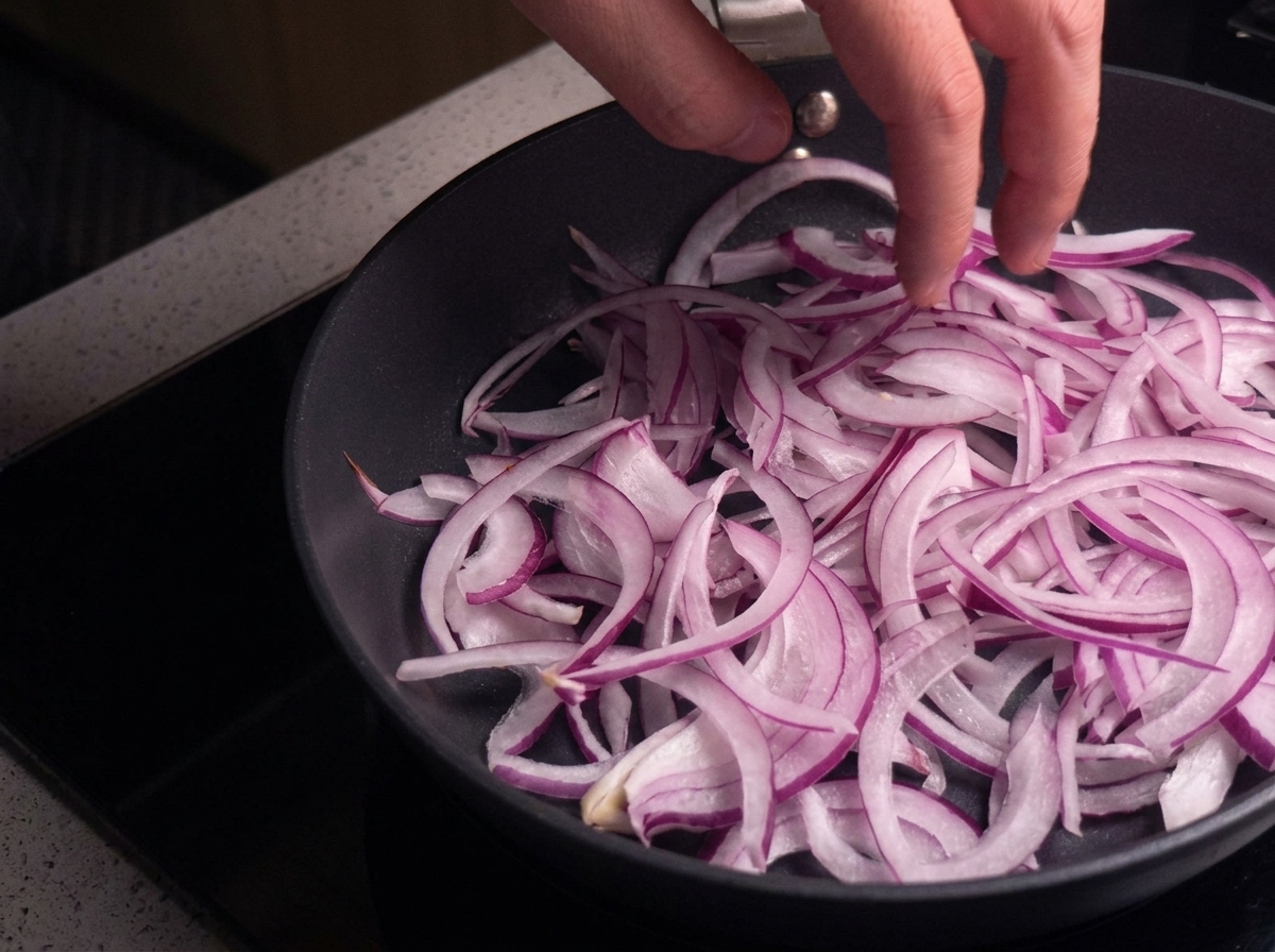 A hand spreading thinly sliced red onions across the surface of a dry black skillet.