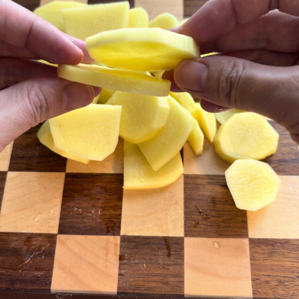 Peeled potatoes being carefully sliced into rounds on a wooden cutting board.