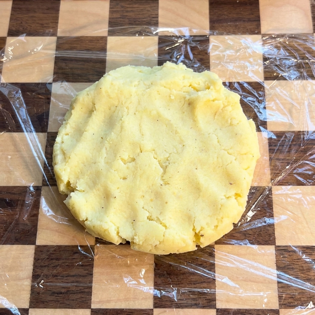 A flat, circular potato patty resting on a checkered cutting board covered with plastic wrap.