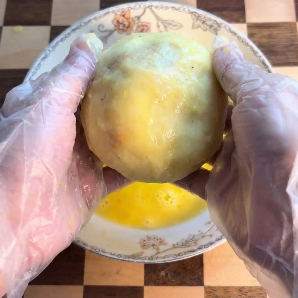 A potato ball being dipped and rolled in a shallow bowl containing yellow whisked egg.