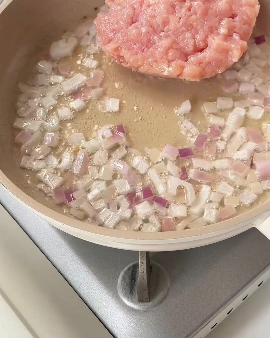 A heap of raw minced chicken placed into a pan with sautéed red onions.