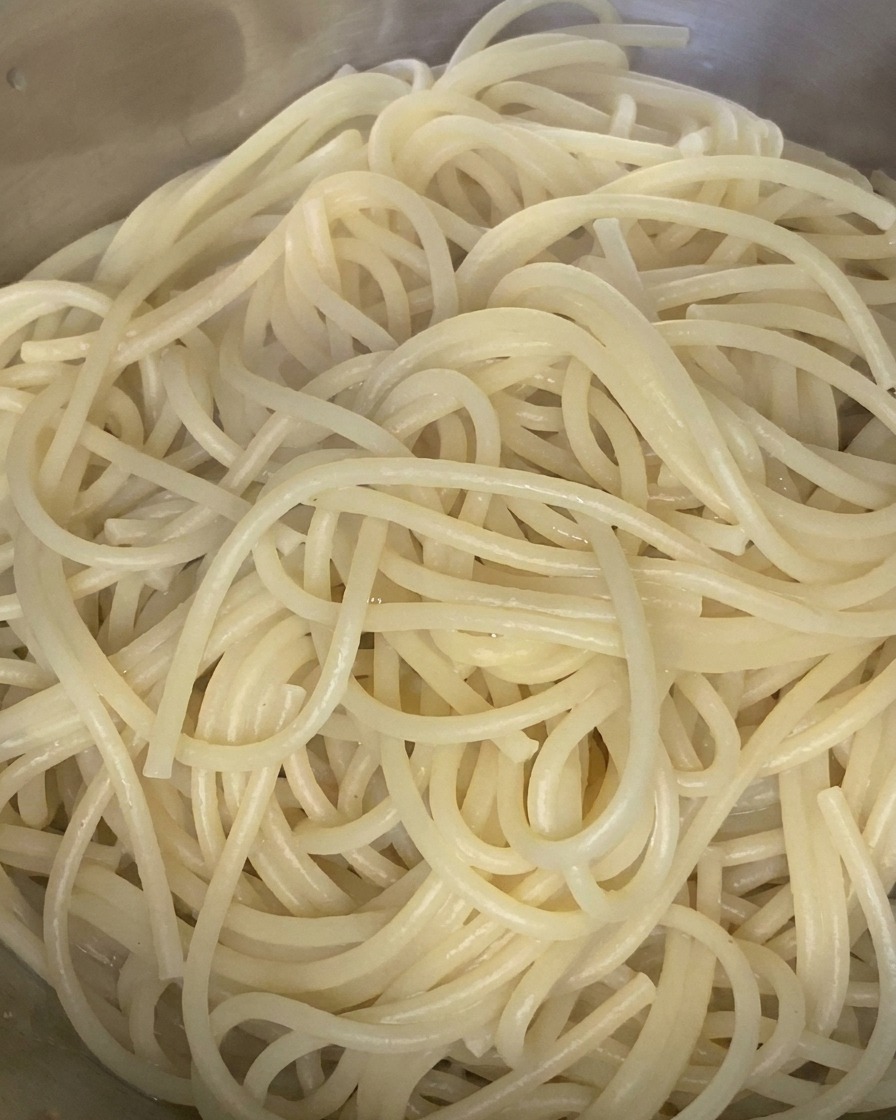 Cooked spaghetti noodles being tossed with a drizzle of olive oil in a stainless steel mixing bowl.