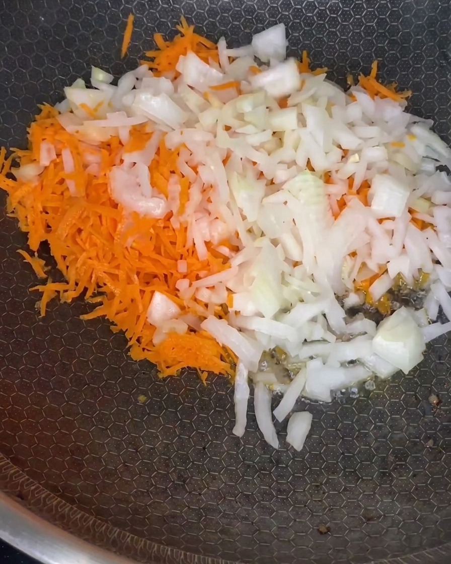 A mix of grated carrots and diced onions softening in a pan over medium heat.