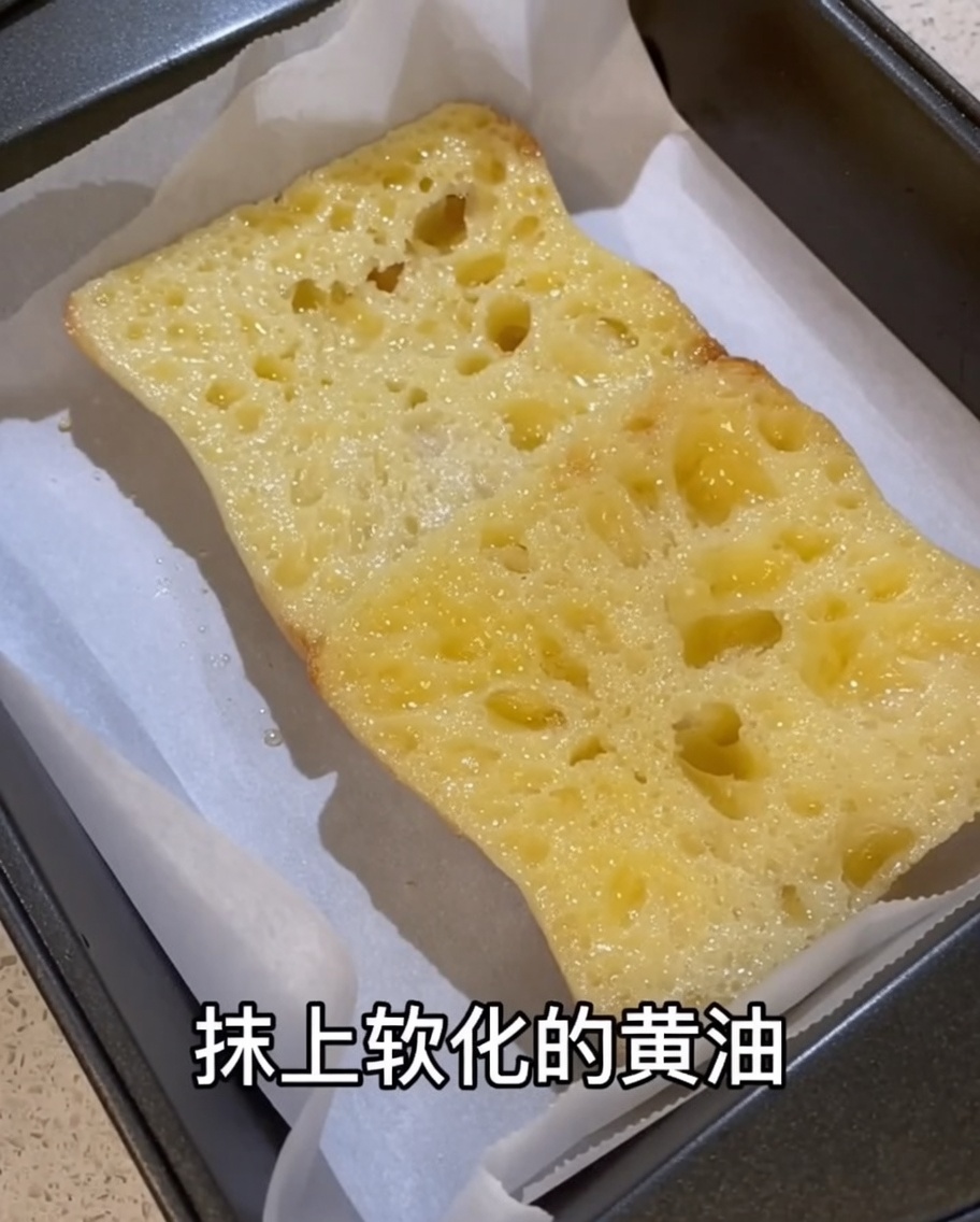 Softened butter being spread onto the surface of a garlic-rubbed ciabatta slice in a parchment-lined baking tray.