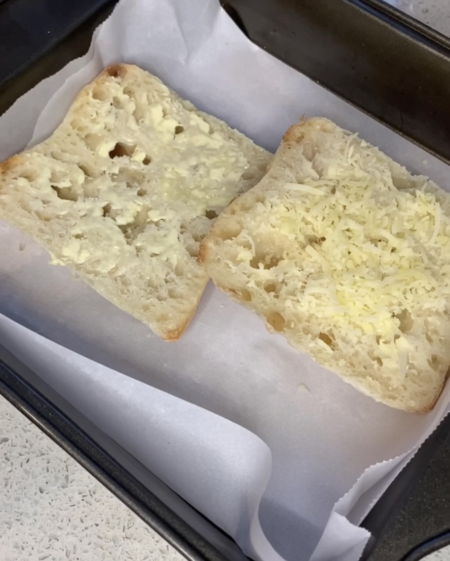 Shredded Parmesan cheese being sprinkled over buttered ciabatta slices on a baking sheet.