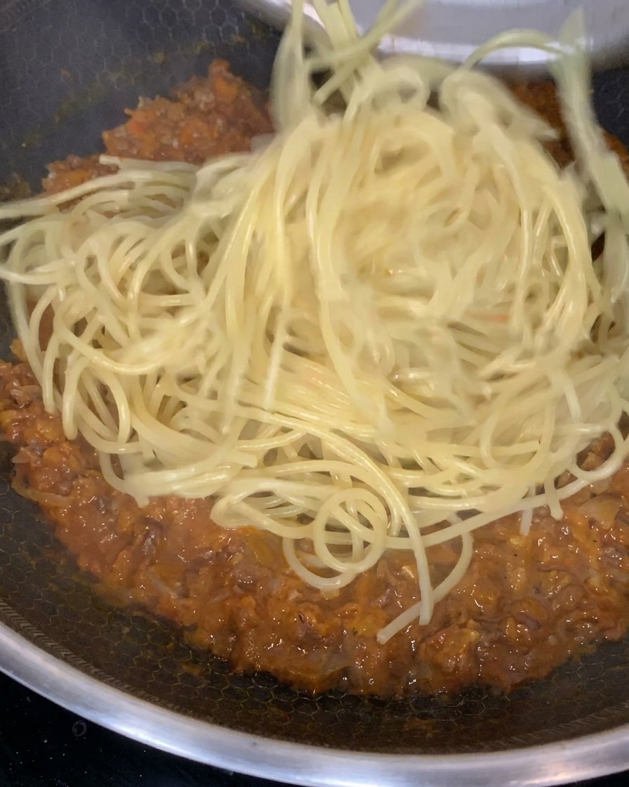 Cooked pasta being tossed and mixed into a rich, thick meat sauce in a large frying pan.