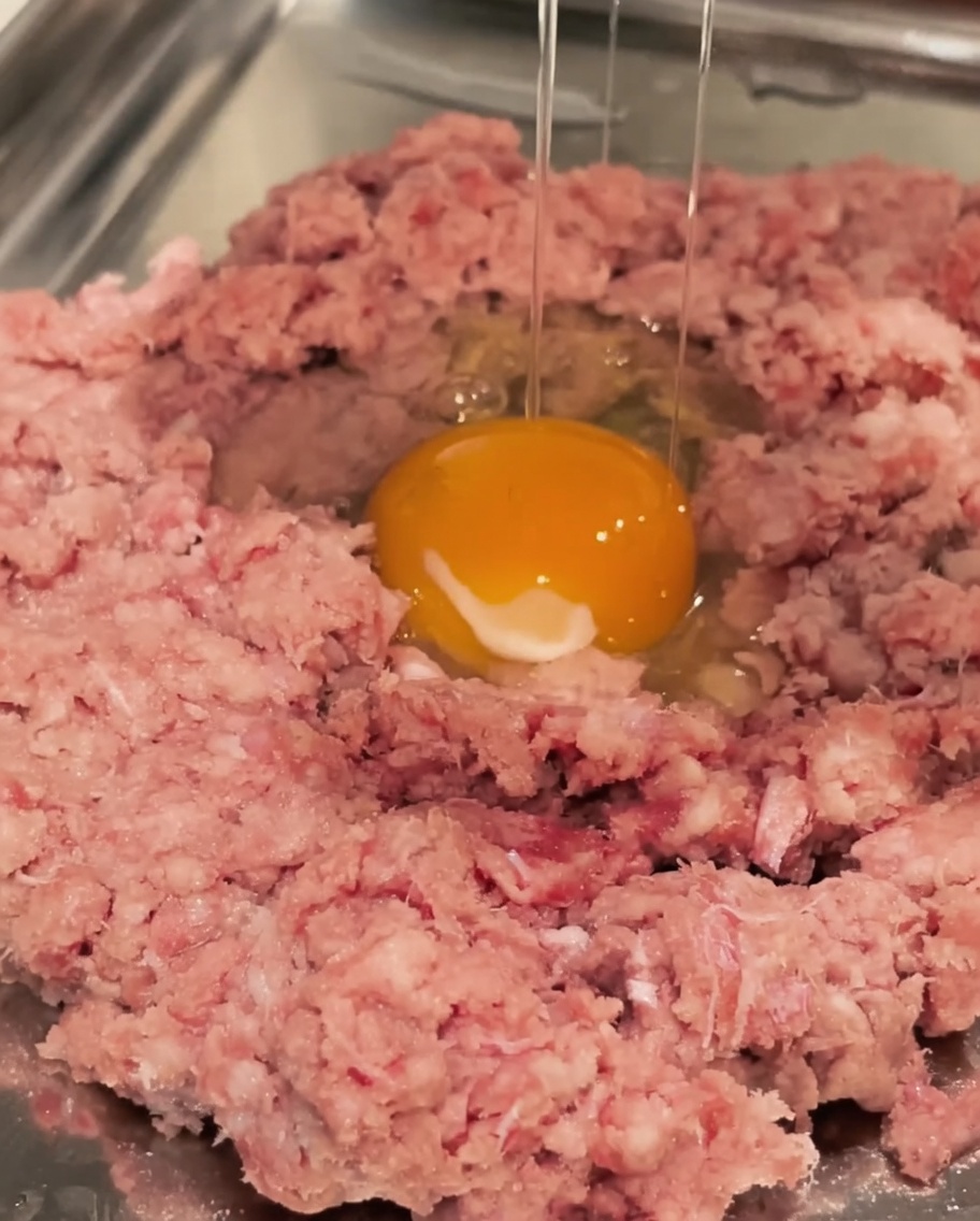 A raw egg being cracked into a bowl of ground beef.