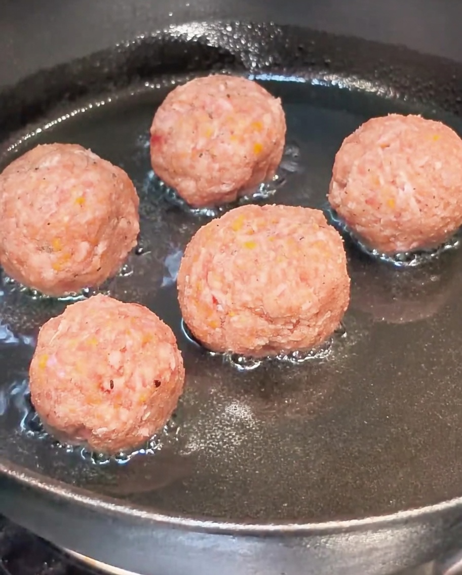 Raw meatballs browning in a hot oiled pan.