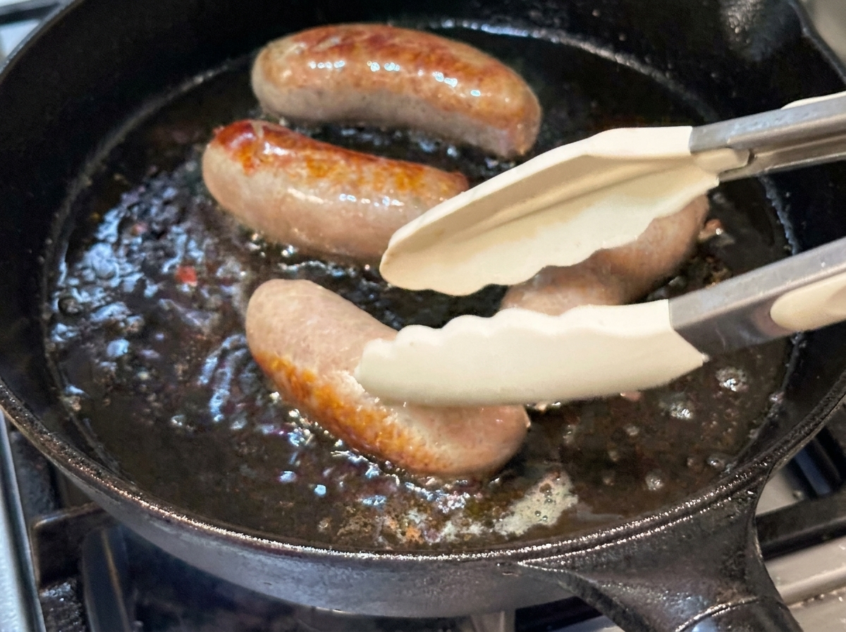 Whole sausages being turned with silicone tongs in a hot cast iron pan.