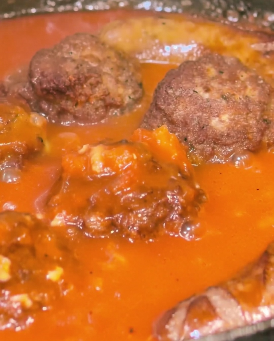 A close-up of a pan containing meatballs and sausages being basted with a thick, red tomato sauce.