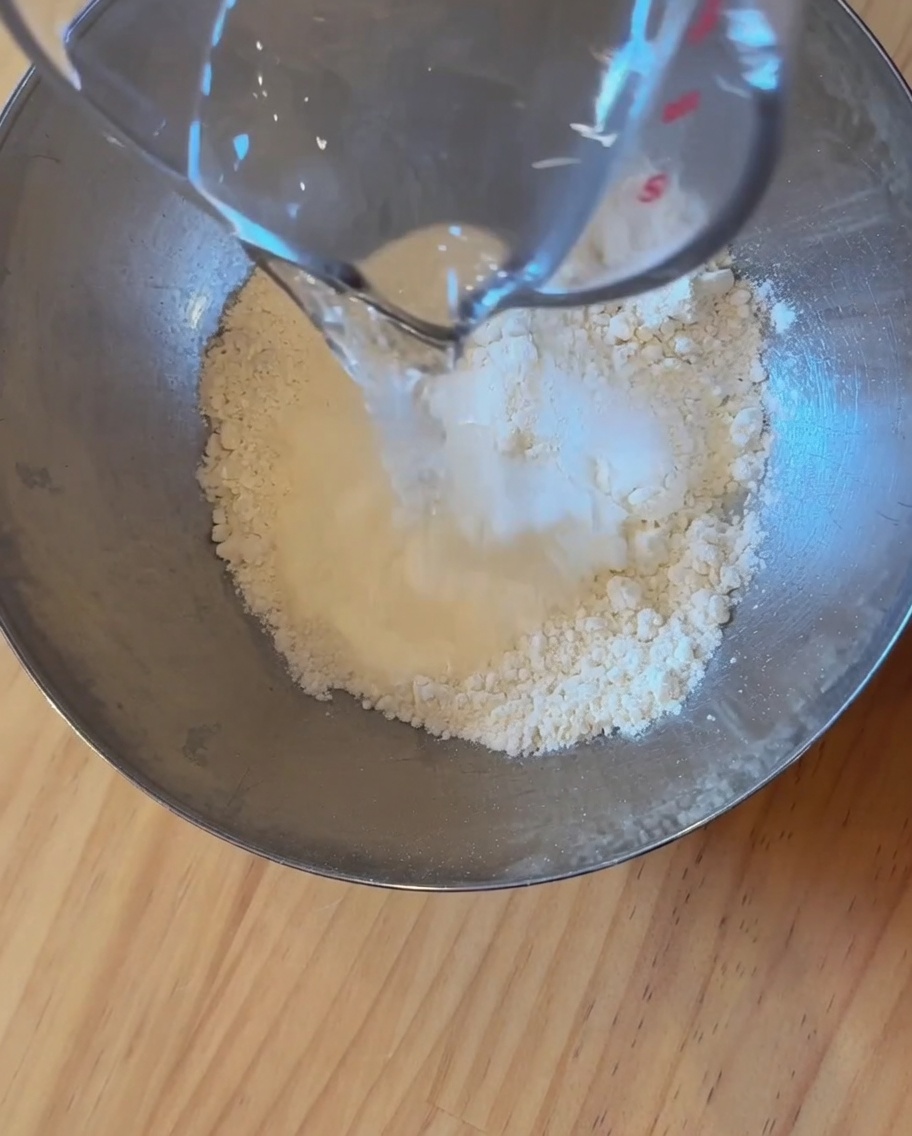 A close-up view of flour, water, and yeast being mixed together in a stainless steel bowl to form a dough.