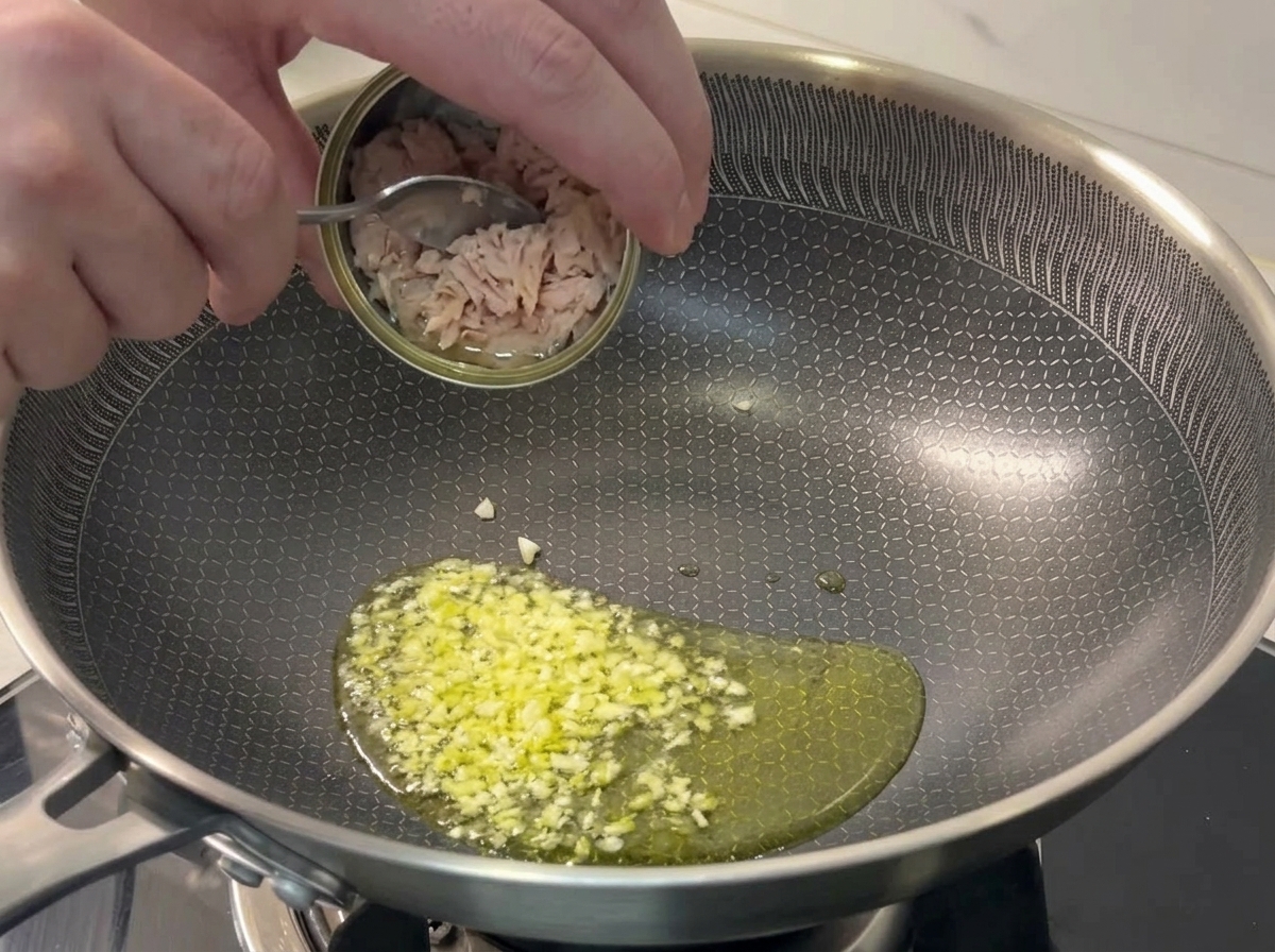 Spooning flaky tuna chunks from a small tin into a pan of sizzling minced garlic.