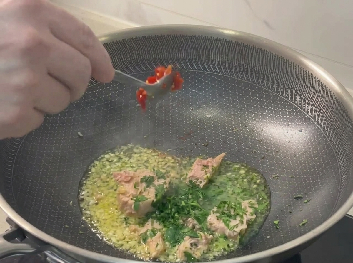 A metal spoon dropping vibrant chopped red chilies into the pan containing tuna, garlic, and parsley.