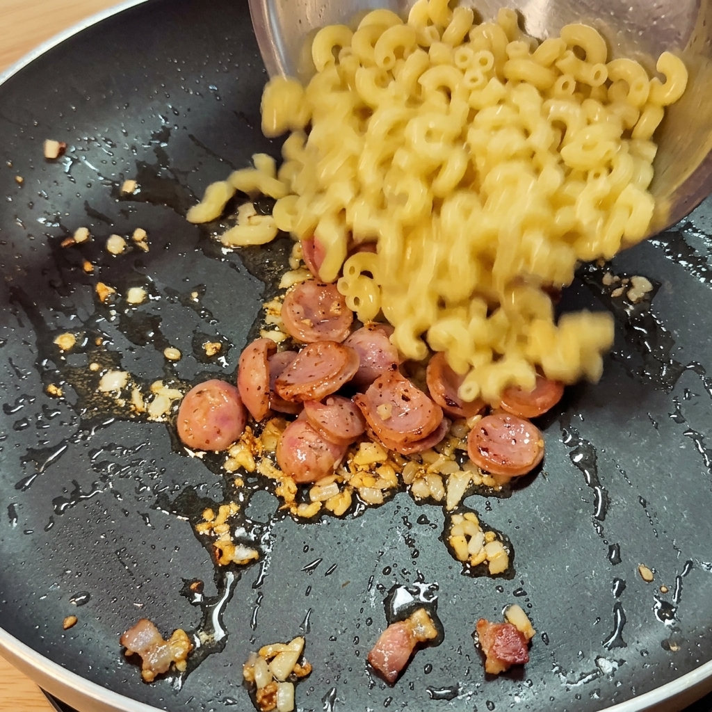 Cooked macaroni being poured into a pan containing fried sausage slices, aromatics, and spicy sauce.