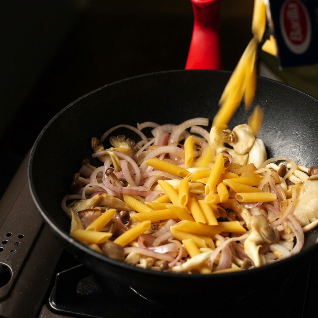 Dry penne pasta pouring from a box into a black pan with sautéed sliced mushrooms and onions.