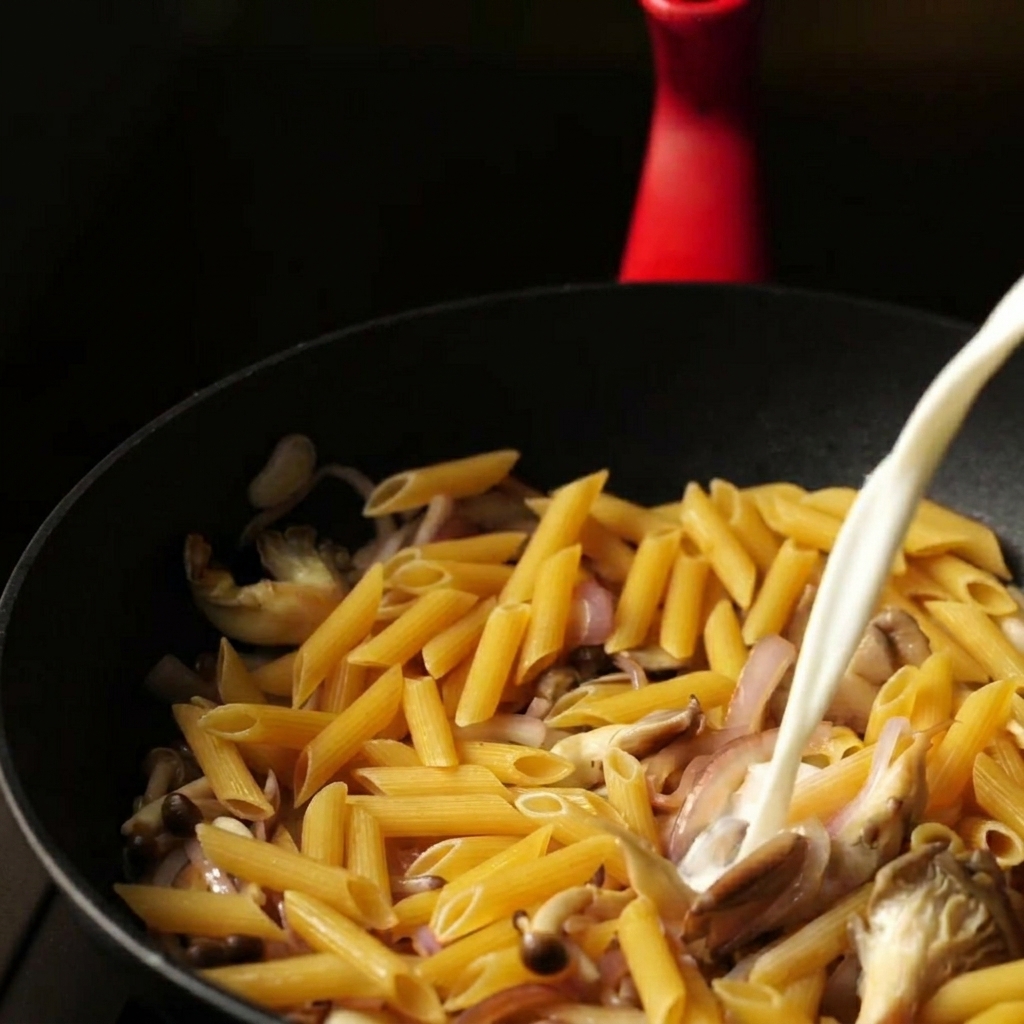 A steady stream of a milk and water mixture being poured into a pan over dry penne and sautéed vegetables.