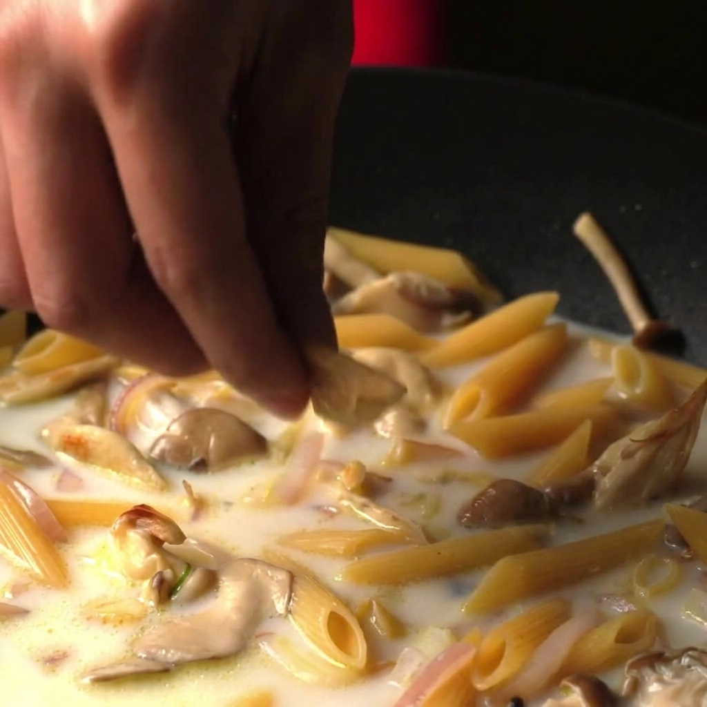 Fingers dropping a piece of bouillon into a pan filled with penne pasta simmering in a creamy white broth.