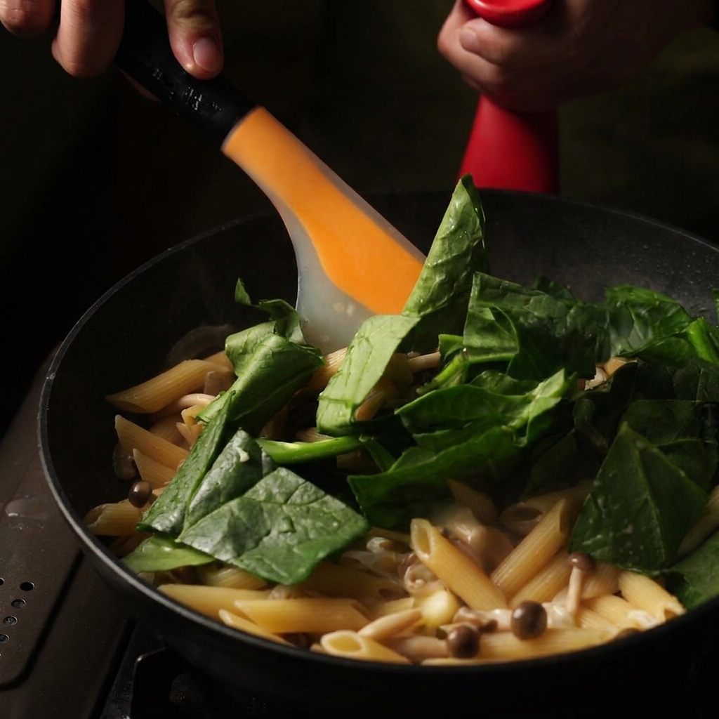 An orange spatula stirring fresh green spinach leaves and butter into a pan of creamy cooked penne pasta.