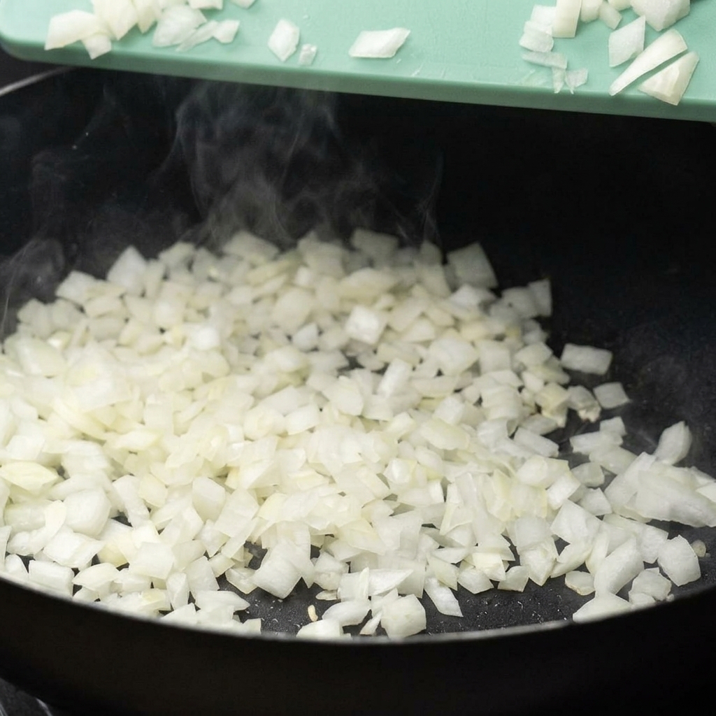 Diced white onions being scraped from a green cutting board into a steaming hot black pan.