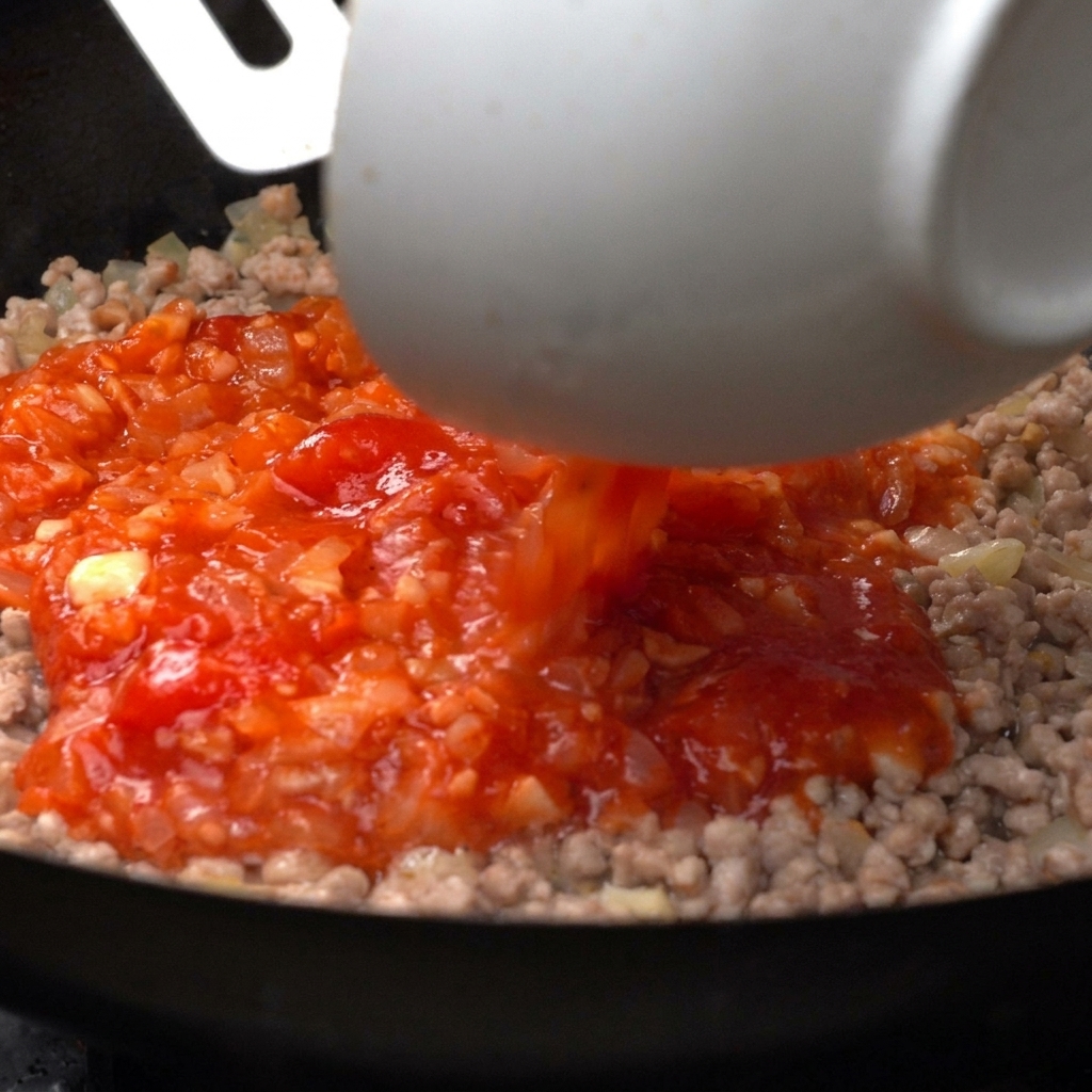 Vibrant crushed tomatoes being poured from a white bowl into a pan of cooked minced meat.