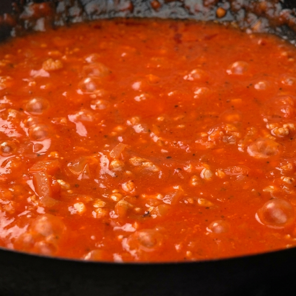 Cooked paccheri pasta being stirred into a thick, vibrant red tomato meat sauce in a pan.