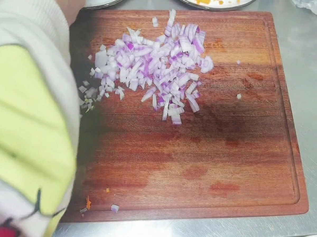 A persons arm visible while chopping a red onion into small dice on a wooden cutting board.