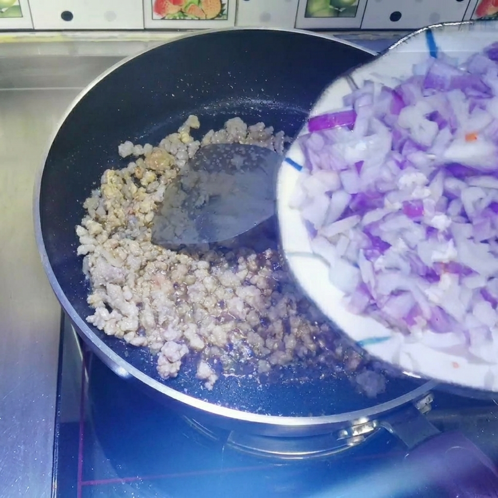 Diced red onions being scraped from a white plate into a non-stick pan containing cooked minced meat.
