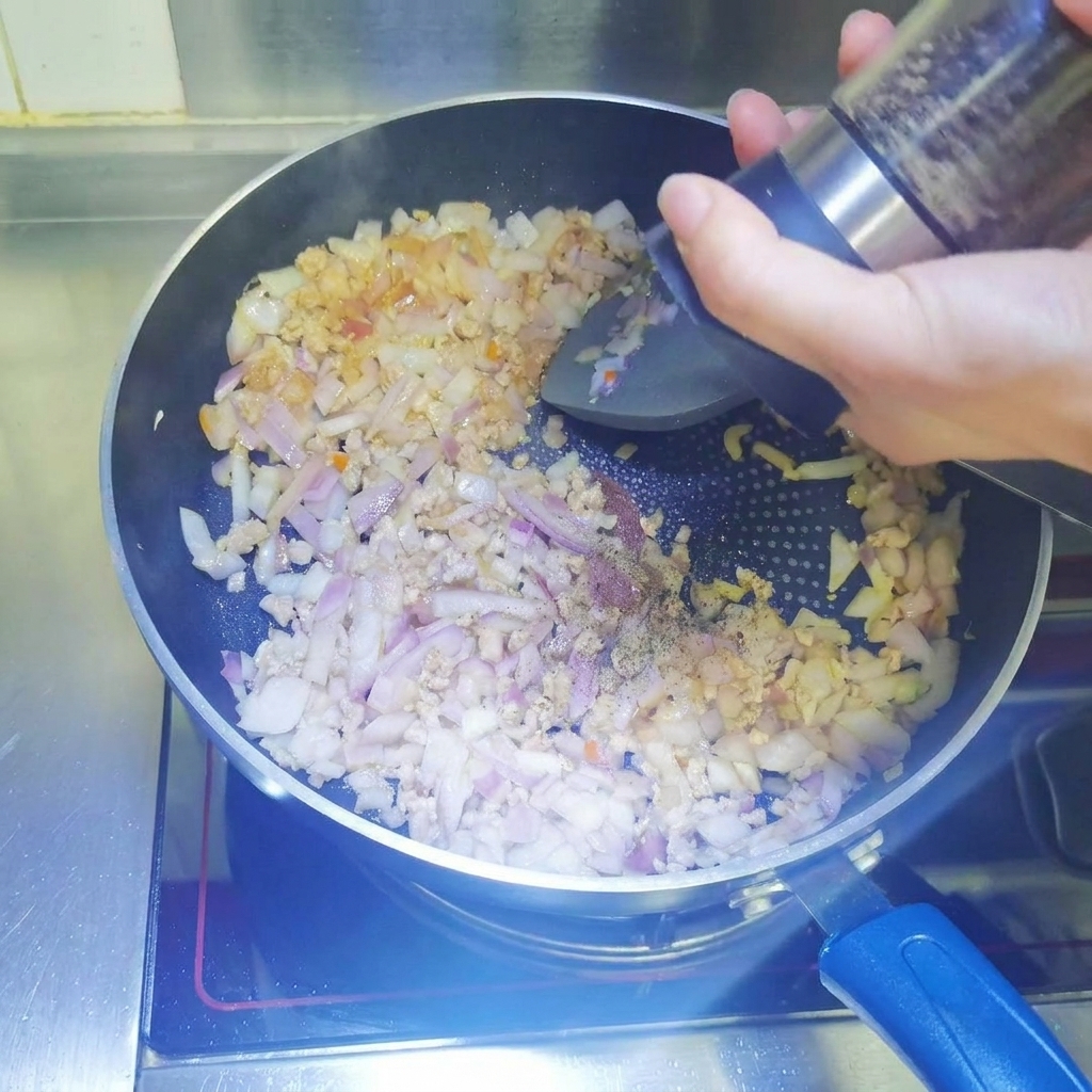 Black pepper being freshly ground from a mill into a pan containing browning minced meat and diced onions.
