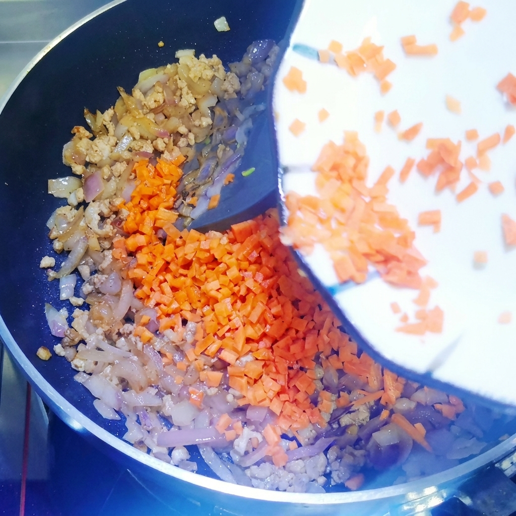 Diced carrots being added to a pan of stir-frying meat and onions, mixed together with a silicone spatula.